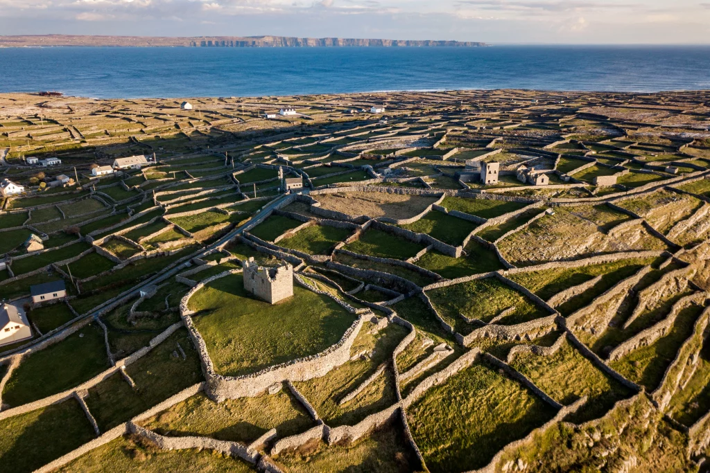 C1 - DJI_0006 Áine Seoighe Aerial view of Inis Oírr, featuring stone walls forming intricate patterns across green fields, with some houses and the sea in the background.