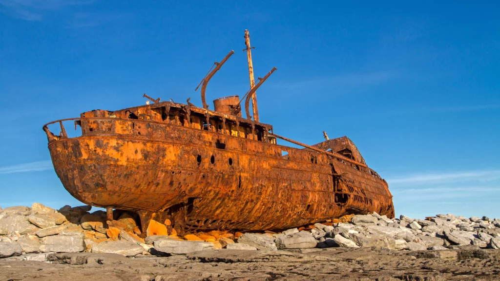 IMG_0676 Áine Seoighe A weathered, rusted shipwreck resting on rocky terrain under a clear blue sky.