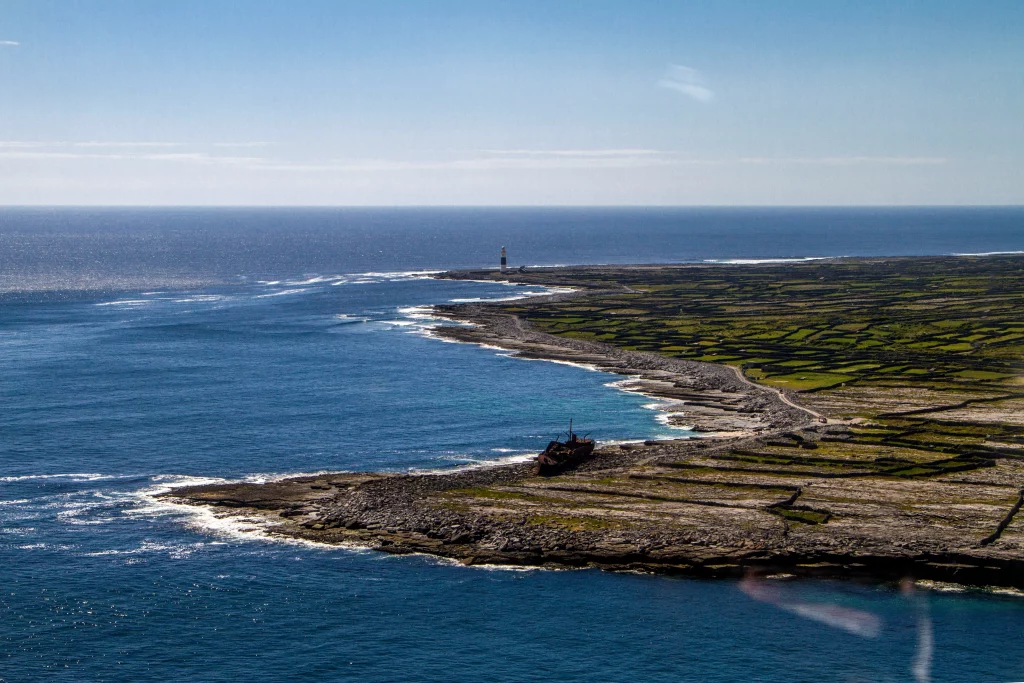IMG_7333 Áine Seoighe Aerial view of Inis Oírr, showcasing a shipwreck on the rocky shoreline, with green fields and a lighthouse in the distance against a blue sea.