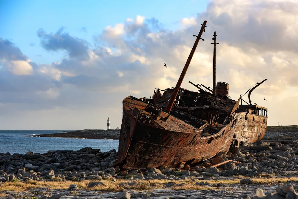 Q2 -PSX_20200605_215649 Áine Seoighe A shipwreck on the rocky shore of Inis Oírr, with a lighthouse visible in the background and a cloudy sky above.