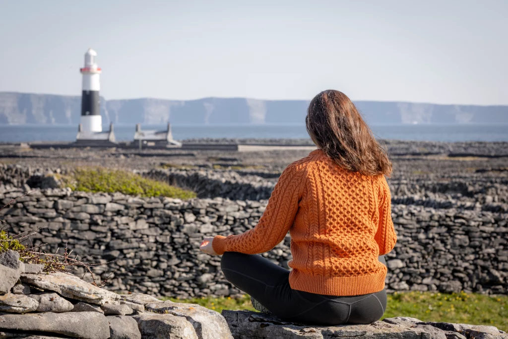 Áine meditates on a stone wall overlooking the sea and lighthouse on Inis Oírr, enjoying the serene landscape of the Aran Islands.