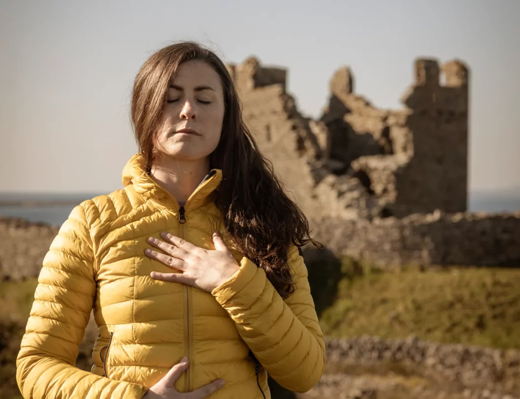 Áine wearing a yellow jacket practices breathwork outdoors on Inis Oírr, with ancient ruins in the background, embodying the spirit of wellness and nature at Éalú Aran retreats.