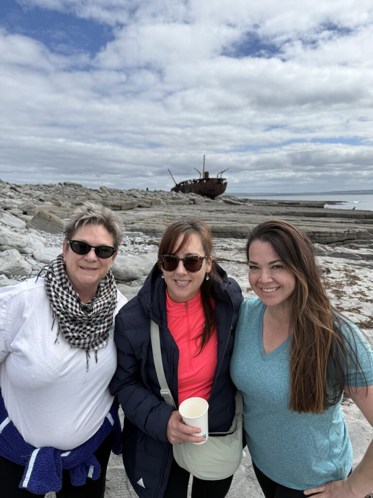 three people standing facing the camera with a shipwreck in the background